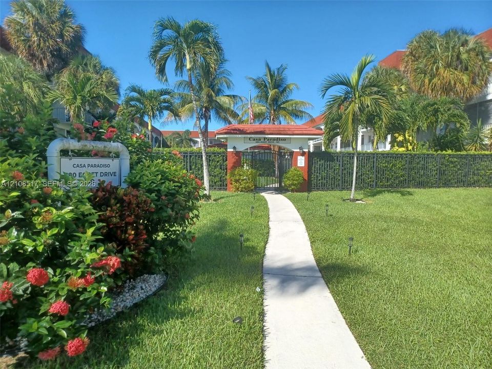 Gated community entrance featuring a red-tiled roof portico, concrete walkway, perimeter black metal fencing, and mature tropical landscaping