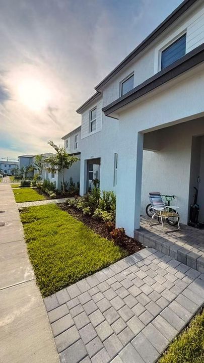 Exterior facade featuring light-toned stucco and horizontal siding, covered entryway with paver flooring, and landscaped front yard with turf and mulch beds