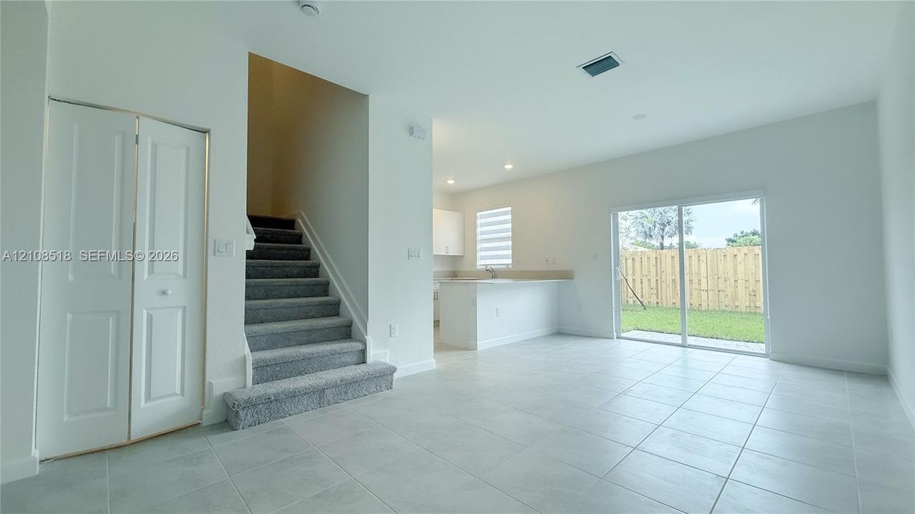 Open-concept interior featuring neutral tile flooring, carpeted staircase with white railings, sliding glass doors, built-in storage closet, and kitchen peninsula