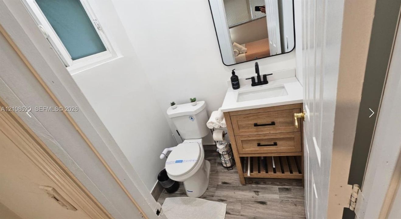 Bathroom featuring a wood-finish vanity with a white countertop and rectangular sink, matte black faucet, rectangular mirror with a black frame, and wood-finish flooring