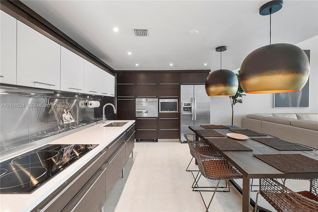 Two-tone cabinetry with white upper and dark wood-finish lower units, stainless steel backsplash, integrated wall oven and microwave, stone-finish countertops, and dual oversized dome pendant lights