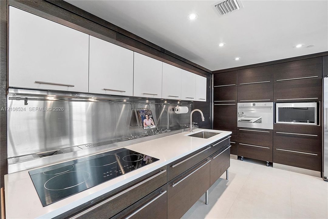 Kitchen featuring white upper cabinetry, dark wood-finish lower cabinetry, metallic backsplash, stone countertops, and built-in stainless steel appliances