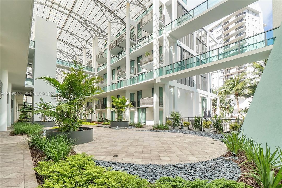 Central atrium featuring a multi-story glass-enclosed canopy, elevated pedestrian bridges, paved hardscaping, and integrated botanical landscaping