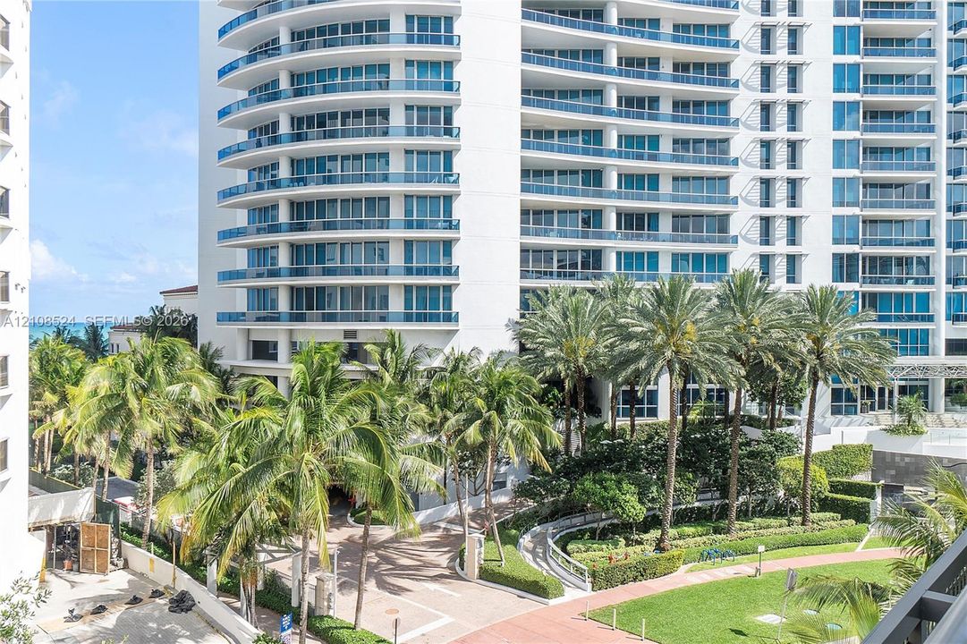 High-rise building exterior featuring curved glass balconies, concrete facade, landscaped grounds with mature palm trees, and paved walkways