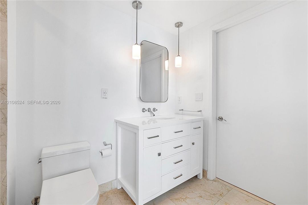 White vanity with a solid-surface countertop and integrated sink, complemented by dual hanging pendant lights, a rounded-corner rectangular mirror, and polished metal hardware