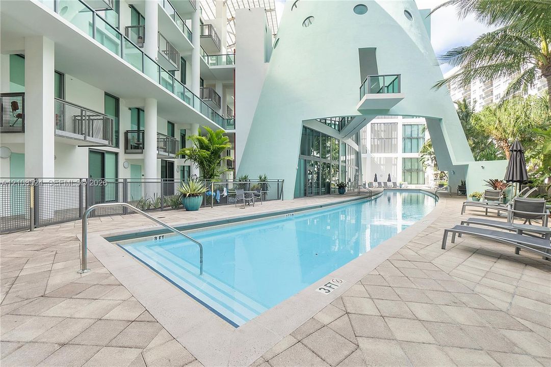 In-ground swimming pool with stepped entry, expansive stone-paver decking, and prominent architectural sculptural facade featuring metal railings and glass balcony enclosures