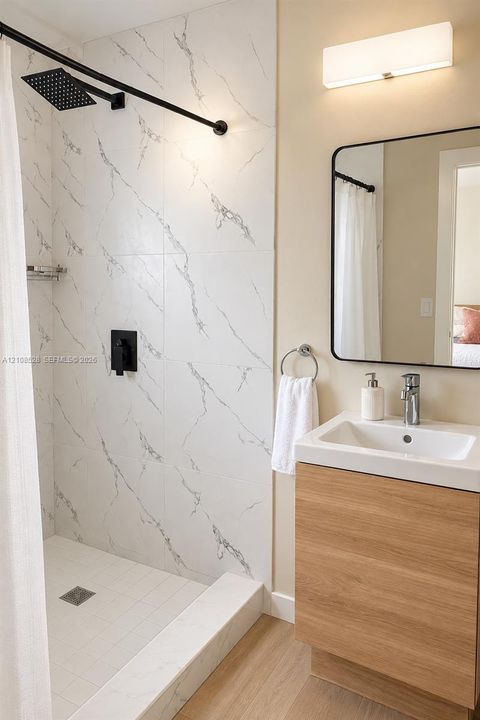 Walk-in shower featuring large-format tile with veining, matte black rainfall shower head, wood-finish vanity with integrated sink, rectangular wall mirror with black frame, and wall-mounted light fixture