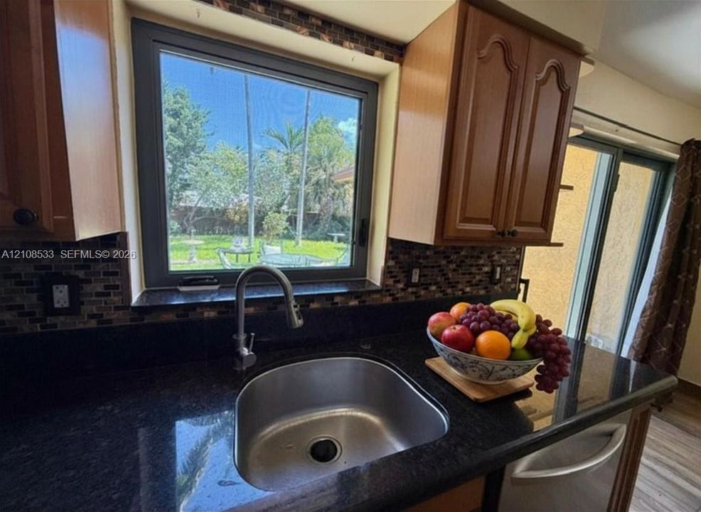Kitchen featuring dark stone countertops, mosaic tile backsplash, wood-finish cabinetry, stainless steel undermount sink with high-arc faucet, and oversized window