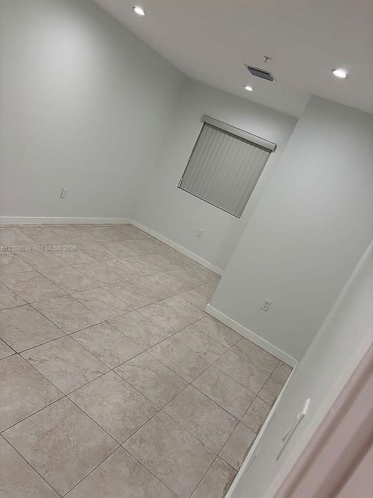 Neutral-toned interior space featuring light-colored tile flooring, recessed ceiling lighting, and a single window equipped with vertical blinds