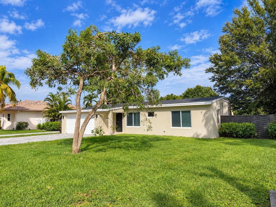 Single-story exterior featuring light-toned stucco siding, a two-car garage, mature landscaping, and an expansive lawn
