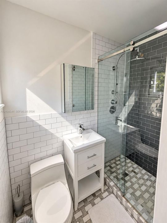 Bathroom featuring white subway tile wainscoting, a white pedestal-style vanity with metal hardware, a glass-enclosed shower with charcoal subway tile surround, multi-tone mosaic floor tiling, and a recessed mirrored medicine cabinet