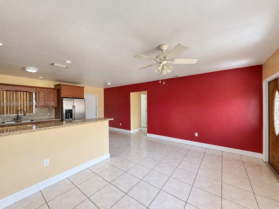 Open-concept interior featuring light-toned tile flooring, kitchen with granite countertops and wood cabinetry, ceiling fan, and wooden entry door with an oval glass insert