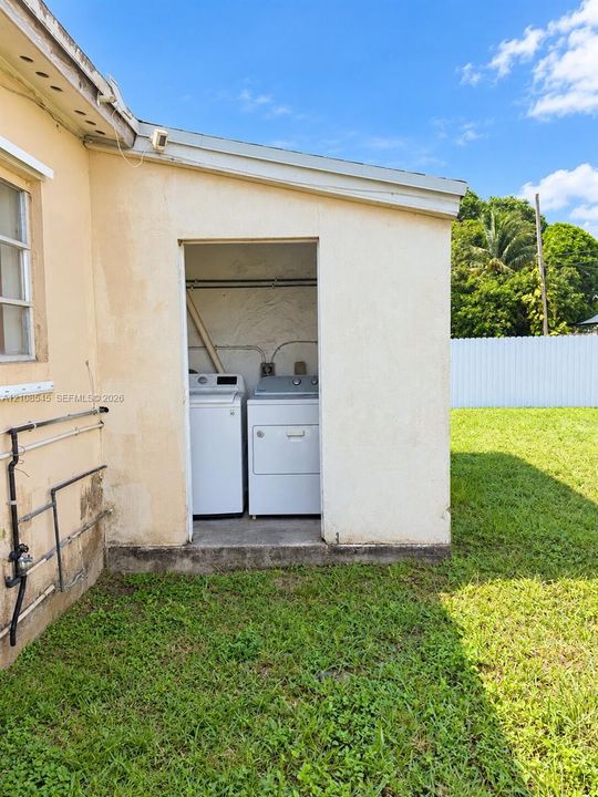 Exterior laundry alcove featuring a concrete foundation, integrated plumbing hookups, and a white vinyl privacy fence bordering the rear lawn