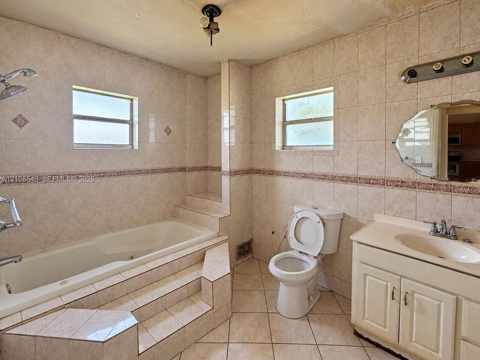 Bathroom featuring full-height ceramic wall tiling, a raised jetted tub with stepped tile surround, a built-in vanity with an integrated sink, and dual windows