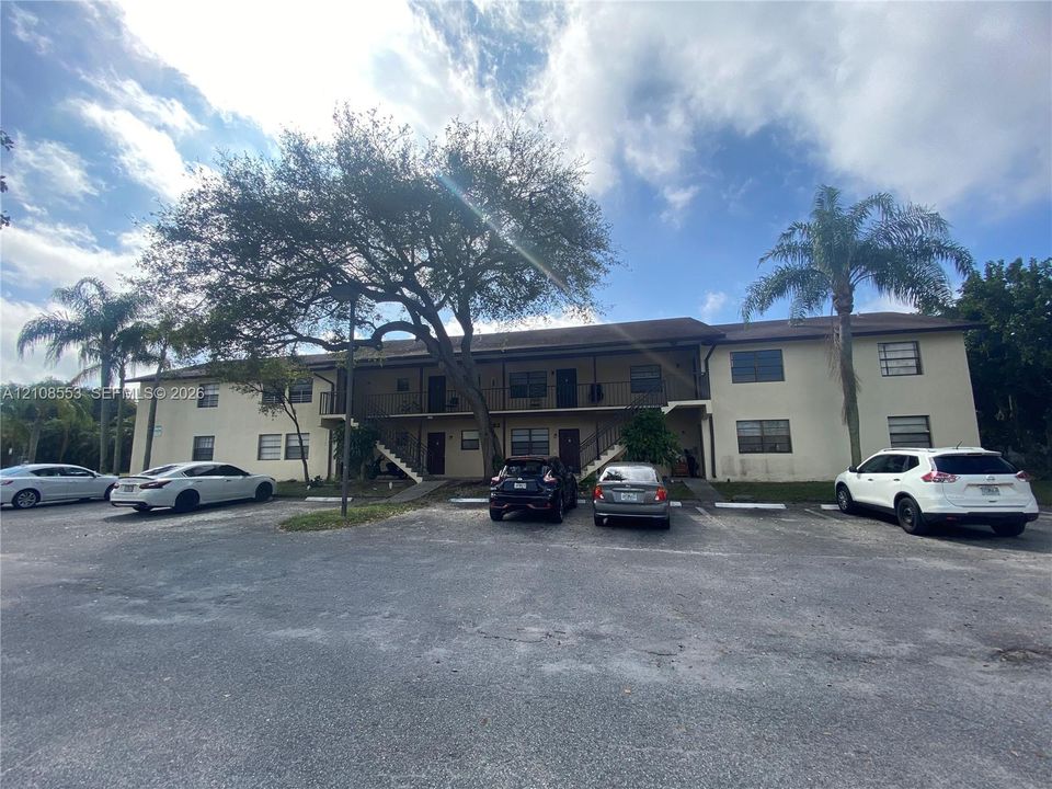 Two-story exterior featuring a neutral stucco finish, covered balcony corridors, mature shade trees, and an asphalt parking lot