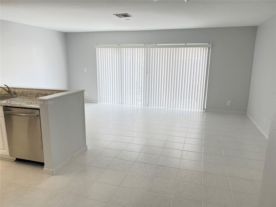 Neutral-toned interior featuring ceramic tile flooring, kitchen peninsula with stone-finish countertop, stainless steel dishwasher, and sliding glass doors with vertical blinds