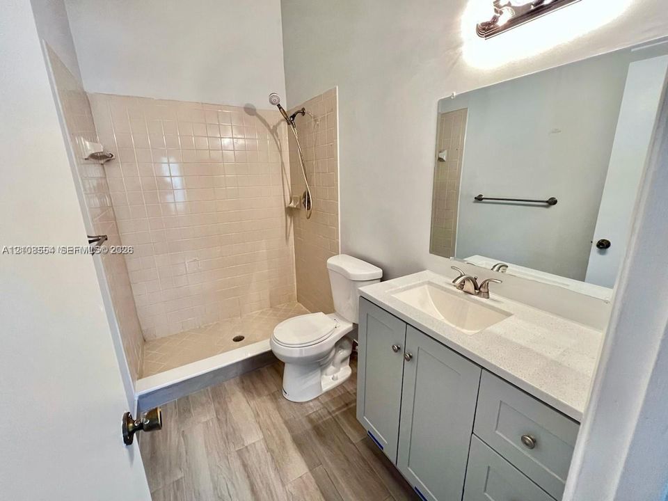 Bathroom featuring a walk-in shower with tile surround, wood-finish flooring, shaker-style vanity cabinet with a stone-composite countertop, undermount sink, and wall-mounted vanity lighting