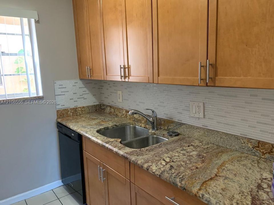 Kitchen featuring warm-toned wood cabinetry with bar-pull hardware, stone-surface countertops, a double-basin stainless steel sink, a mosaic-tile backsplash, and a built-in black dishwasher