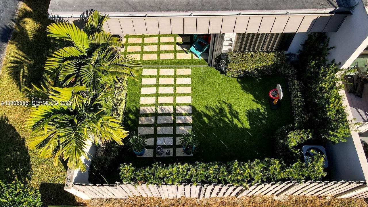 Enclosed backyard featuring a paver stone walkway, artificial turf lawn, mature palm trees, perimeter hedging, and wood-style privacy fencing