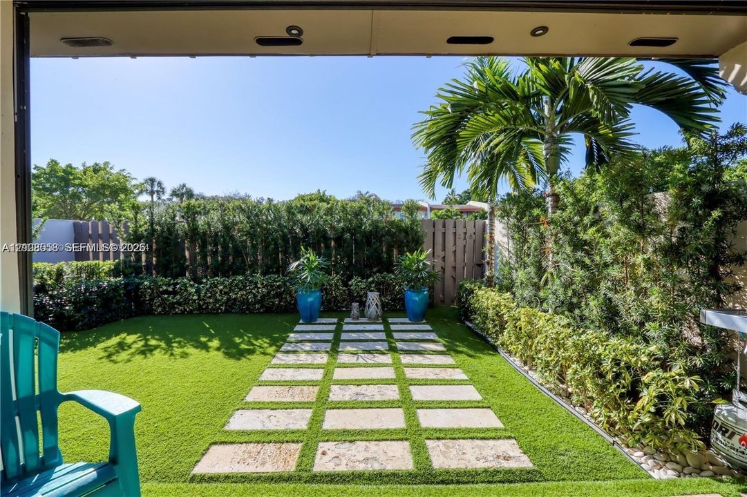Enclosed backyard featuring synthetic turf, stone paver pathway, mature perimeter hedge privacy screen, and recessed soffit lighting