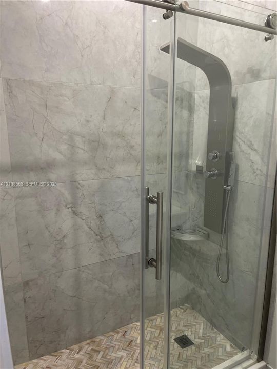 Walk-in shower featuring large-format stone-pattern wall tile, herringbone-pattern stone tile flooring, a glass sliding door with metal hardware, and a multi-function shower panel system