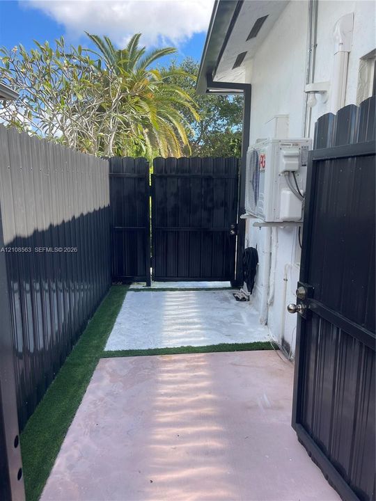 Side exterior space featuring privacy metal fencing, a gated entry, and a paved walkway bordered by synthetic turf
