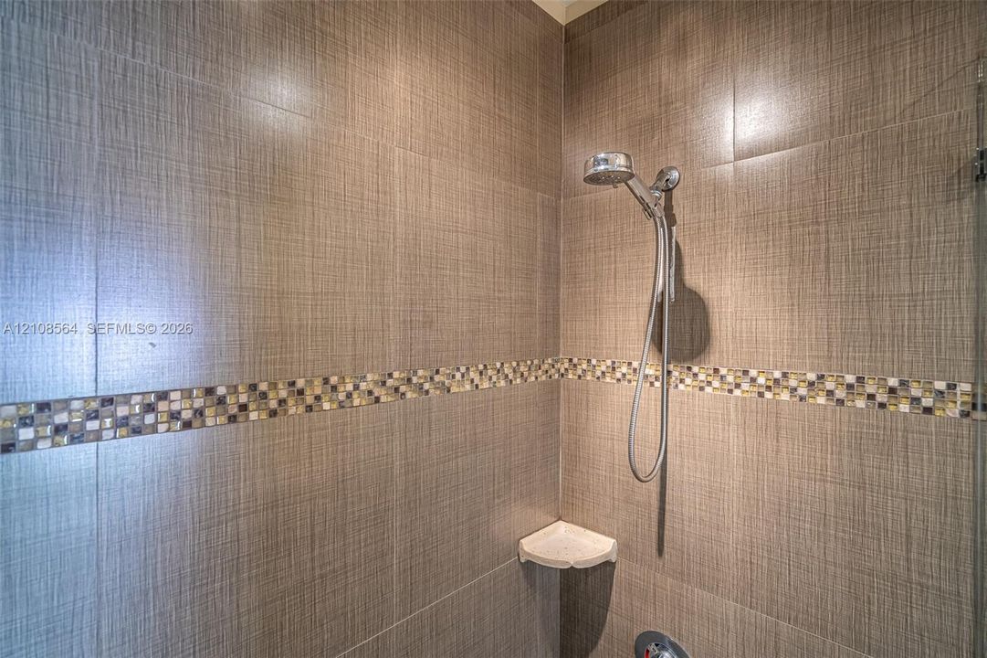 Shower enclosure featuring large-format textured wall tile, decorative mosaic tile inlay, chrome handheld shower fixture, and a built-in corner shelf