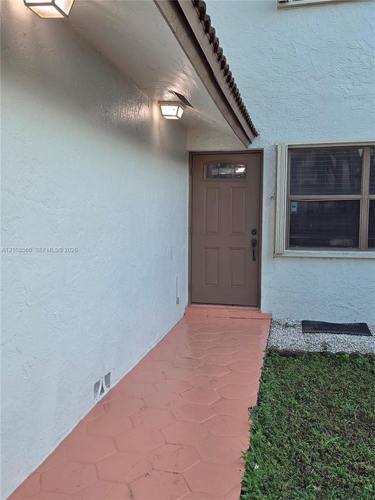 Entrance featuring a solid-panel door with an arched glass inset, hexagonal paver walkway, stucco exterior, and dual wall-mounted outdoor light fixtures