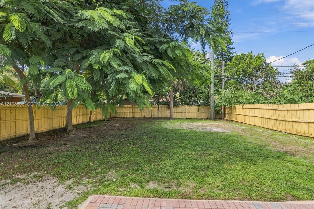 Rear yard featuring a grass lawn, perimeter wood fencing, mature trees, and a paved brick patio