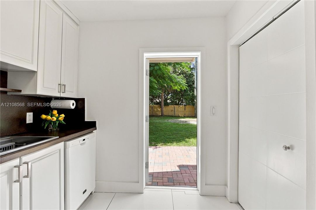 White cabinetry with modern hardware, contrasting dark countertops, built-in dishwasher, large-format tile flooring, and a glass-paneled door opening to a brick paver patio and lawn