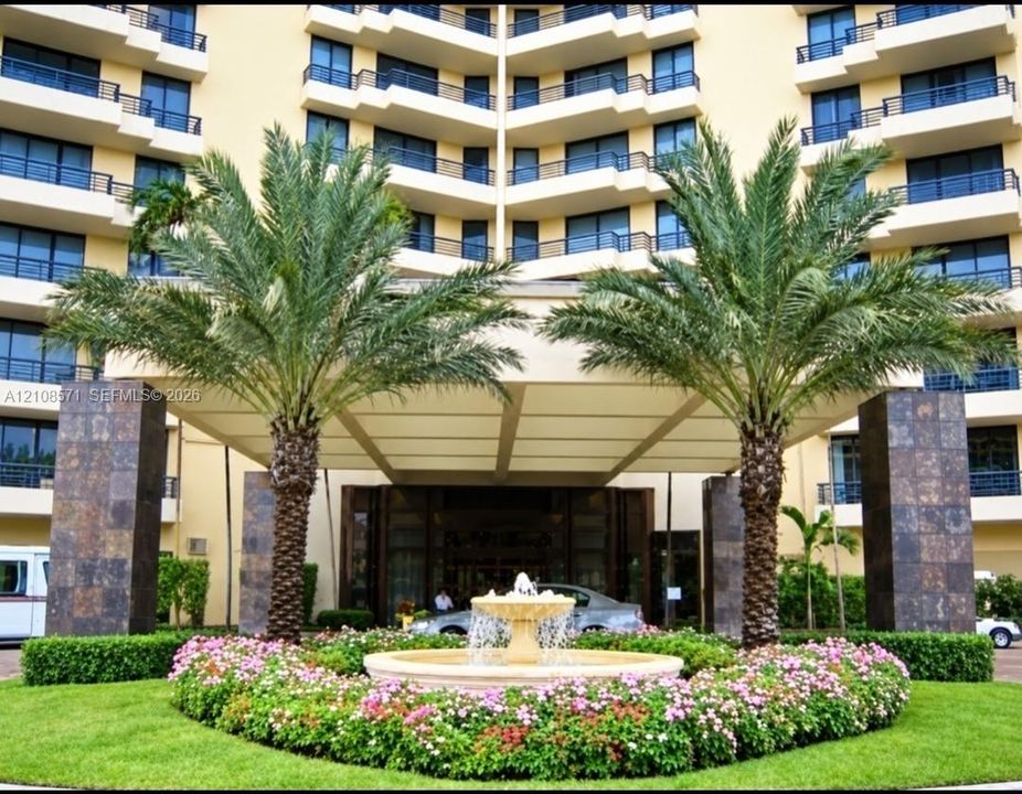 Multi-story building facade featuring a covered porte-cochère, decorative central stone fountain, mature palm trees, and manicured landscaping with flowering perennials