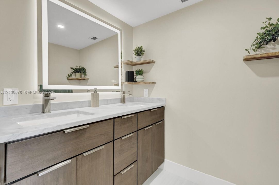 Dual-sink vanity with stone-surfaced countertop, integrated backlit mirror, wood-finish cabinetry, and built-in wall shelving