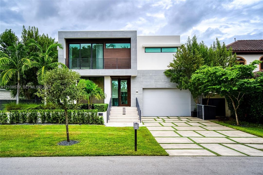 Contemporary multi-level exterior featuring smooth stucco and stone-finish cladding, horizontal wood paneling, metal balcony railing, and a grid-patterned concrete paver driveway