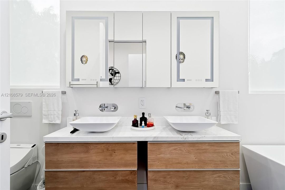 Dual-sink vanity featuring a stone-composite countertop, wood-finish cabinetry, rectangular vessel sinks, wall-mounted faucets, and integrated LED-lit medicine cabinets
