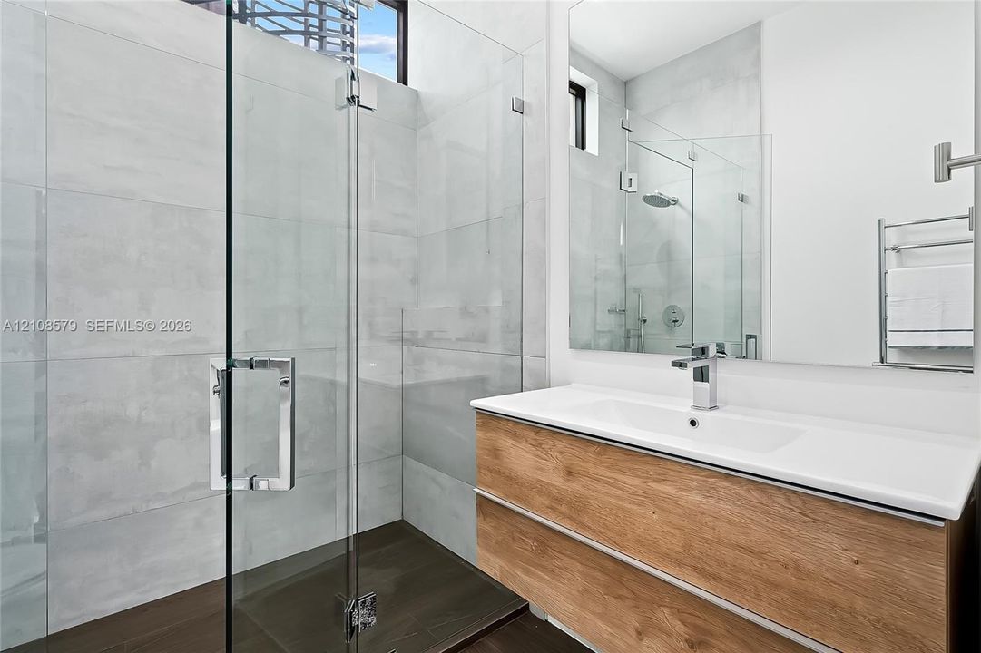 Floating wood-finish vanity with an integrated rectangular sink, large-format tiled walk-in shower with a glass enclosure, wall-mounted heated towel rack, and polished chrome fixtures