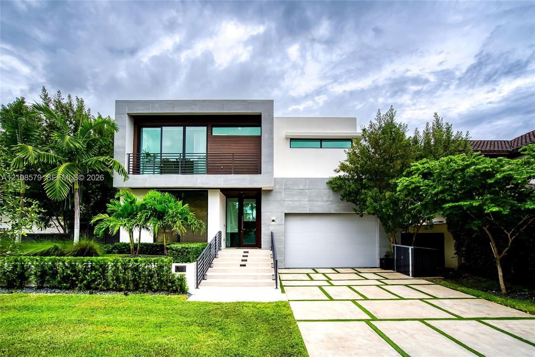 Contemporary two-story exterior featuring a stone-clad facade, wood-panel siding, large-format paver driveway with turf accents, second-story balcony with metal railings, and wide concrete steps leading to the main entrance
