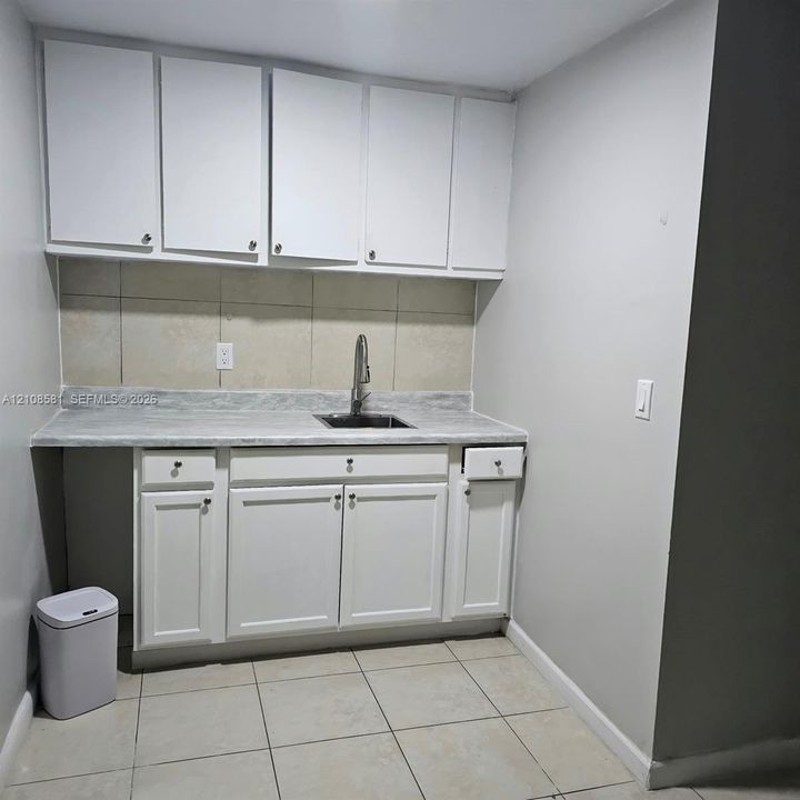 Kitchen area featuring white shaker-style cabinetry, neutral stone-finish countertops, stainless steel undermount sink with high-arc faucet, neutral tile backsplash, and tile flooring