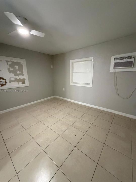 Room featuring large-format neutral tile flooring, a ceiling-mounted light fixture with fan blades, a single-hung window with white trim, and a wall-mounted air conditioning unit