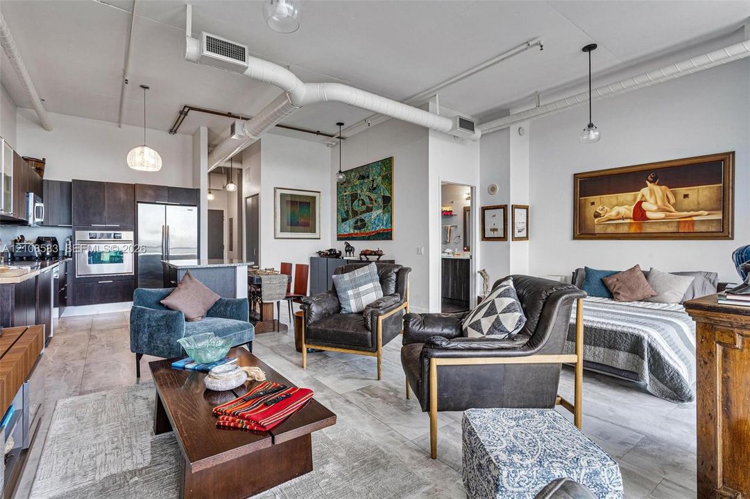 Open-concept industrial-style interior featuring high ceilings with exposed ductwork, dark-finish cabinetry, built-in stainless steel appliances, and large-format tile flooring