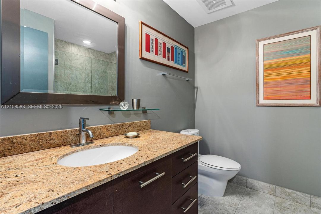 Dark wood-finish vanity with stone-style countertop and undermount sink, complemented by a large framed mirror, glass shelving, and stone-tile flooring