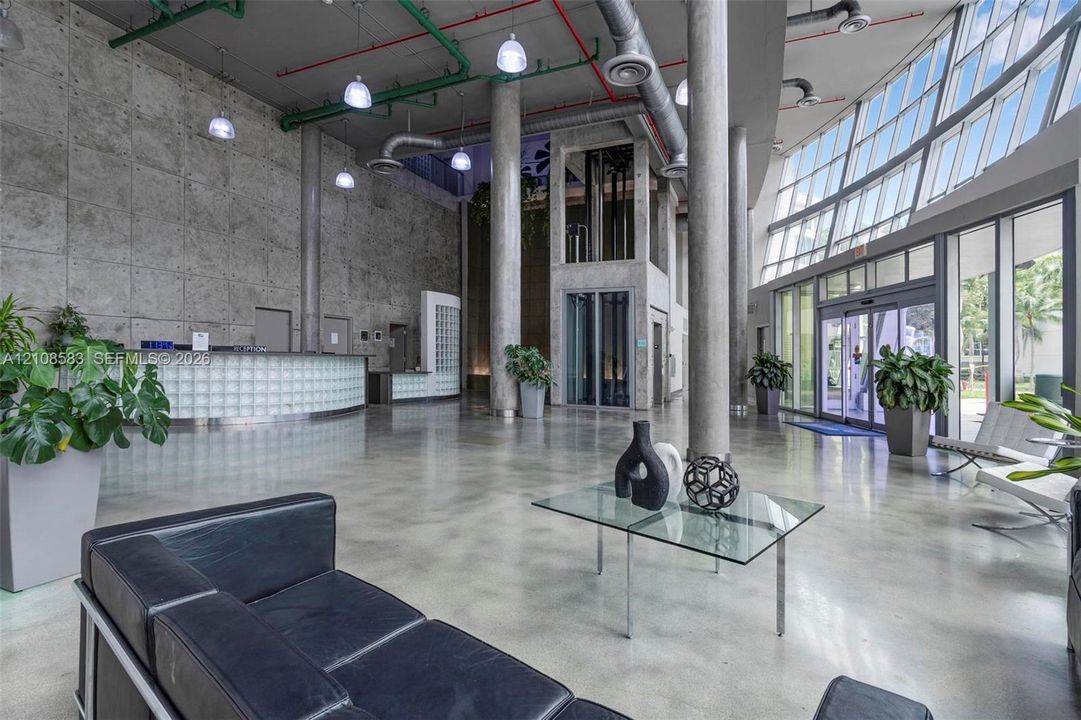 Industrial-style lobby featuring floor-to-ceiling concrete walls, high exposed ceilings, polished concrete flooring, extensive glass curtain walls, and a glass-block reception desk