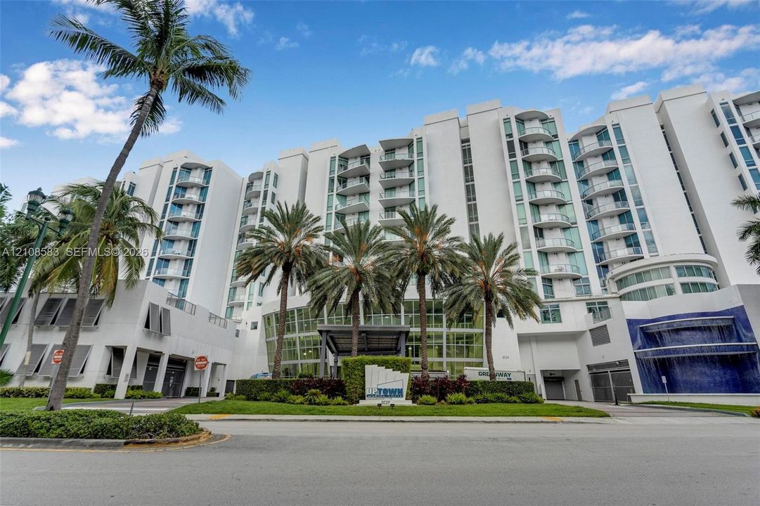 Multi-story high-rise exterior featuring white structural facade, private balconies, glass-paneled window systems, covered entrance, and landscaped grounds with mature palm trees