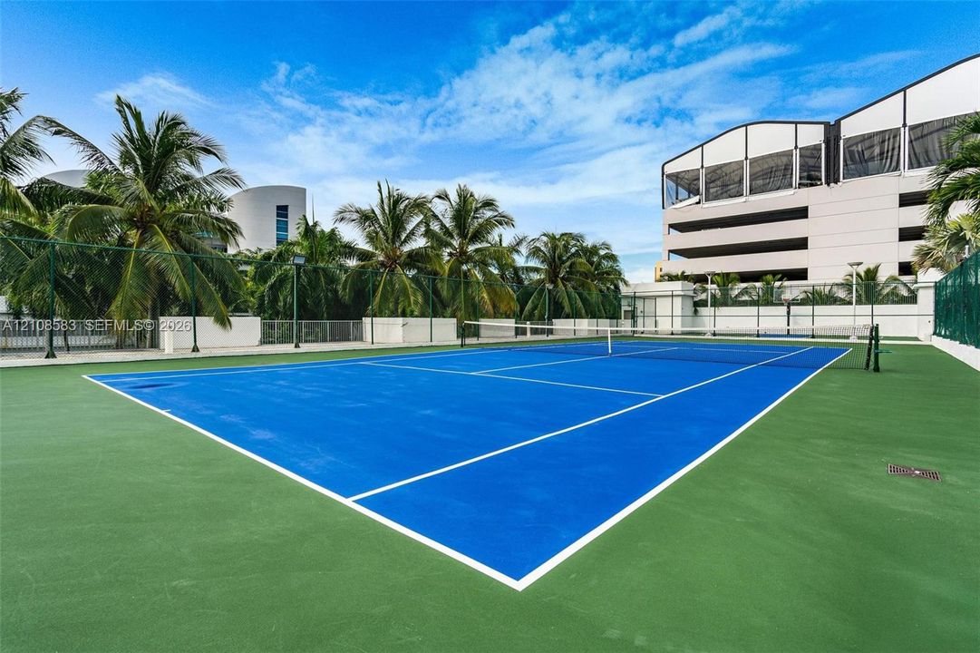Hard-court tennis facility featuring perimeter fencing, pole-mounted lighting, and mature palm trees