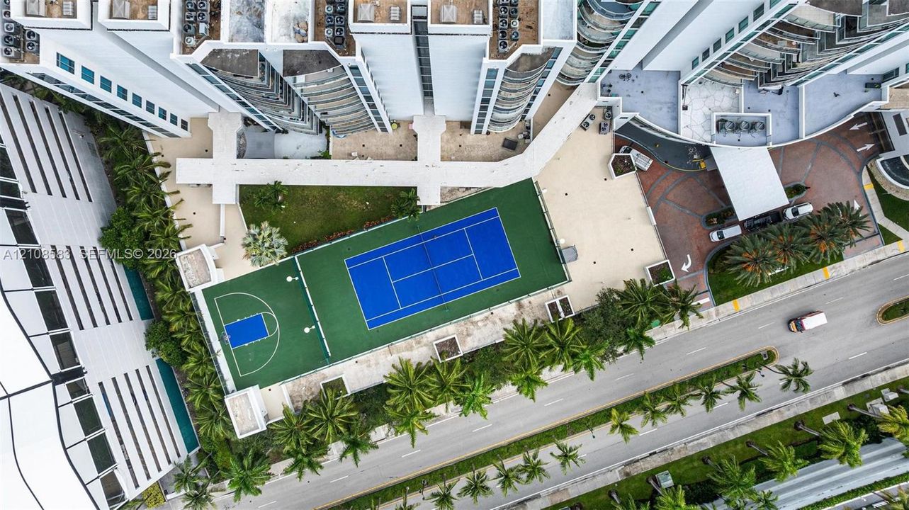 Aerial perspective of the property featuring a multi-sport court with tennis and basketball configurations, paved recreational grounds, and extensive palm tree landscaping
