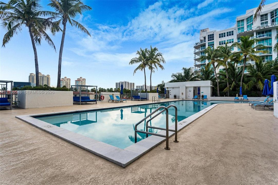 Rectangular outdoor swimming pool featuring a light-colored masonry deck, metal entry railings, concrete perimeter wall, and mature palm trees