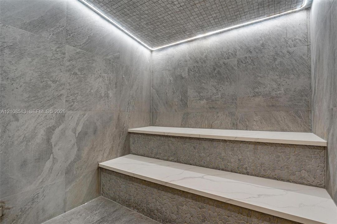 Steam room featuring large-format stone-finish wall and floor tiling, multi-level tiered seating with smooth light-toned surfaces, textured stone-detail risers, and integrated perimeter LED cove lighting