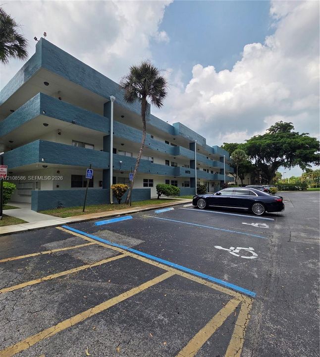 Three-story multi-unit structure featuring a textured blue facade, recessed balconies, and paved surface parking