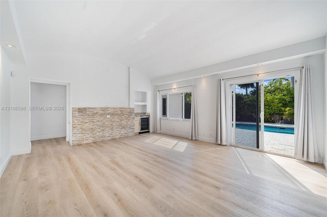 Open-concept interior featuring light wood-finish flooring, vaulted ceilings, a stacked stone accent wall, a built-in beverage refrigerator, and sliding glass doors with exterior pool access