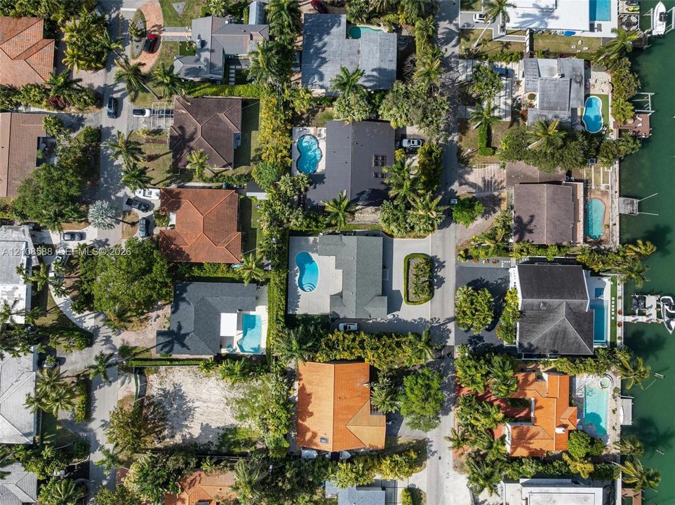 Aerial perspective of a coastal residential neighborhood featuring waterfront properties, private swimming pools, paved driveways, and mature landscaping