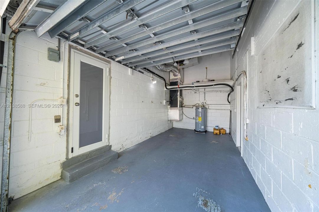 Enclosed garage featuring painted concrete block walls, painted concrete flooring, exposed ceiling joists, a dedicated utility area with a water heater, and a metal-frame exterior entry door with an elevated threshold